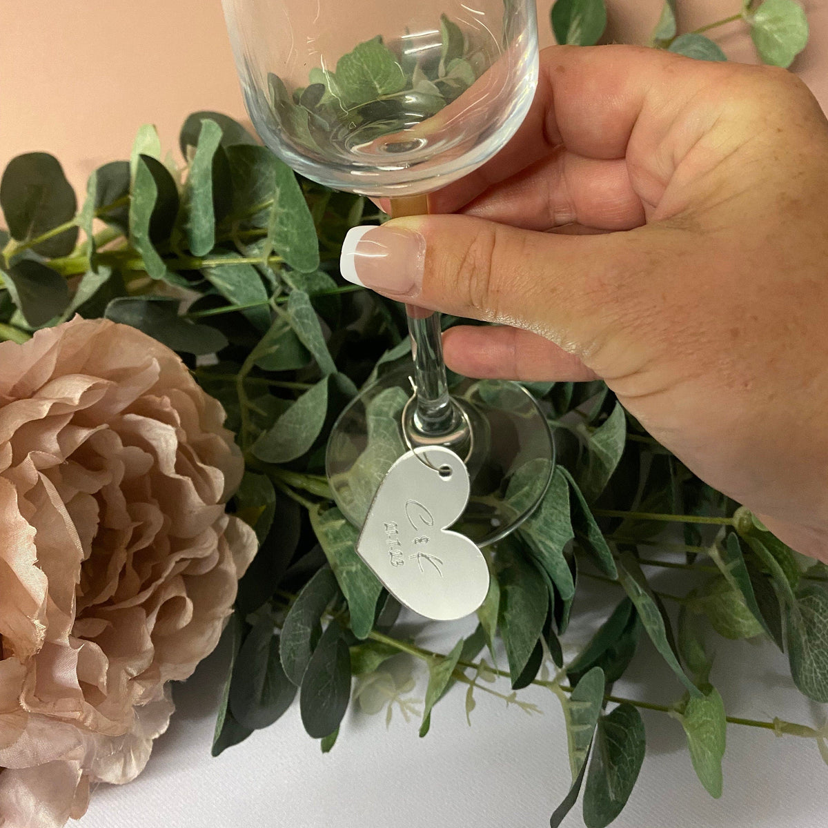 Hand holding a wine glass with a decorative tag next to flowers and greenery on a peach background